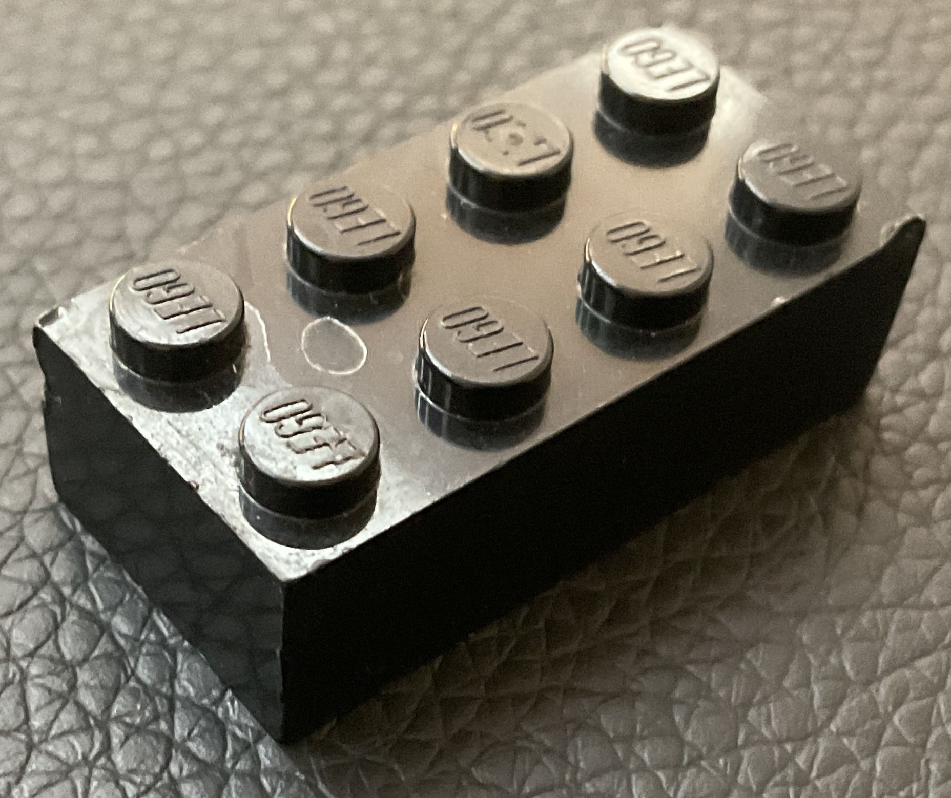 A black 2×4 LEGO brick on leather, eight studs each stamped with the LEGO name