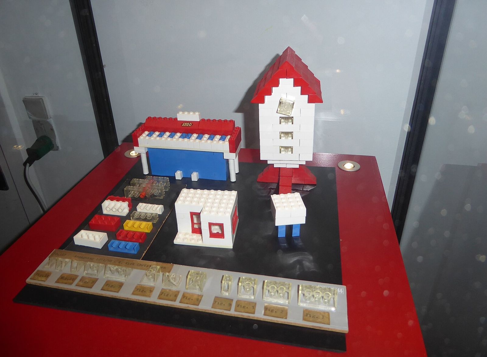 A museum case of early LEGO models: a red and white rocket, small houses, and a row of loose bricks