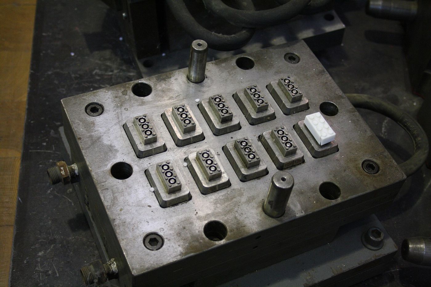 A steel LEGO injection mould with rows of 2×4 brick cavities, one white brick resting on it