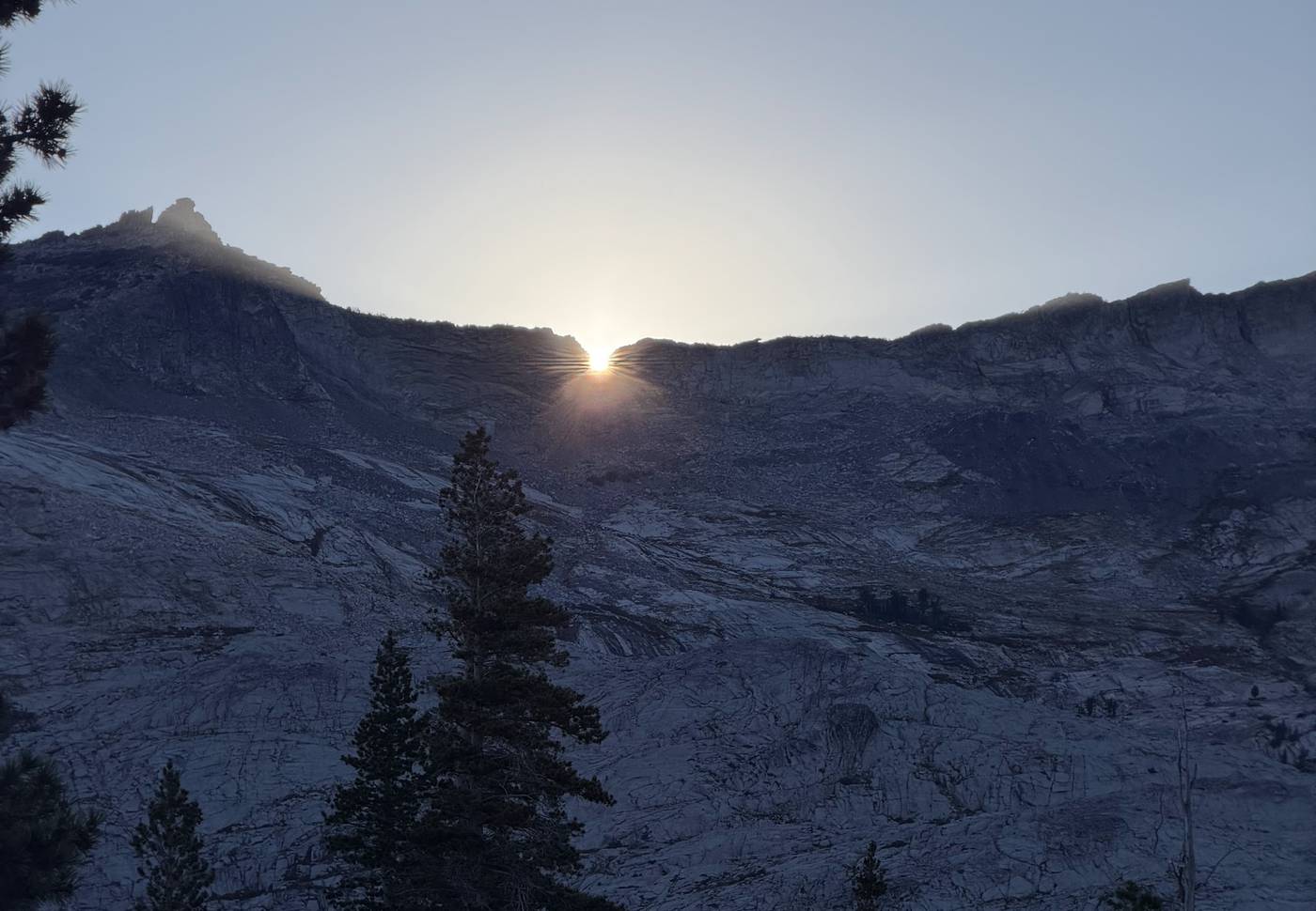 The sun peeks through a notch in a granite ridgeline above a steep slope of pines in Desolation Wilderness