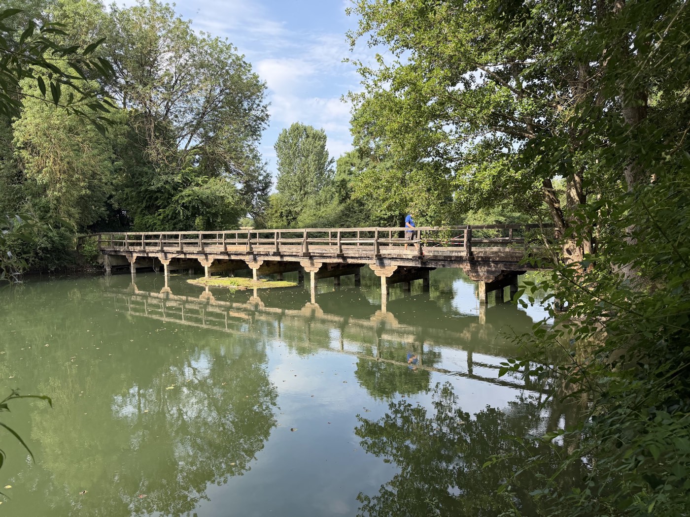 A weathered wooden bridge crossing the still, green Loir, mirrored in the water and framed by trees