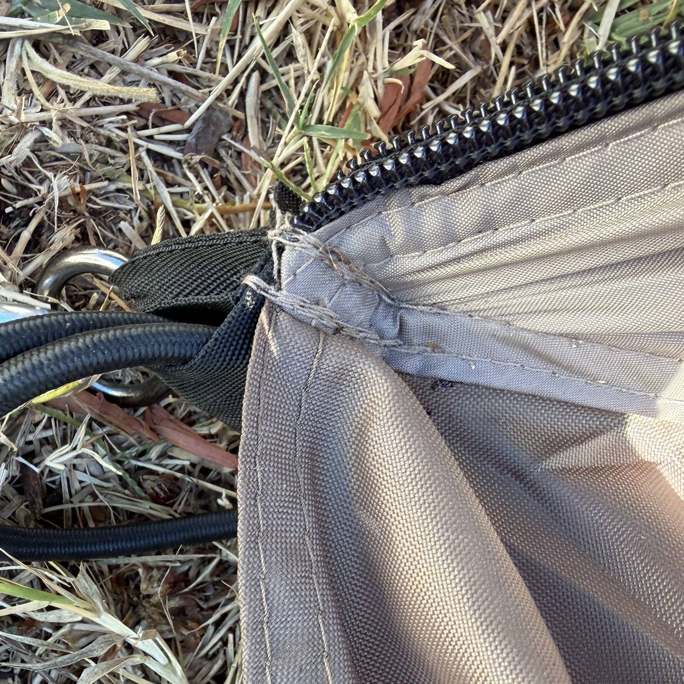 Close-up of uneven hand stitching reattaching a webbing tie-off to the corner of a tan tent, next to a black bungee cord and steel ring